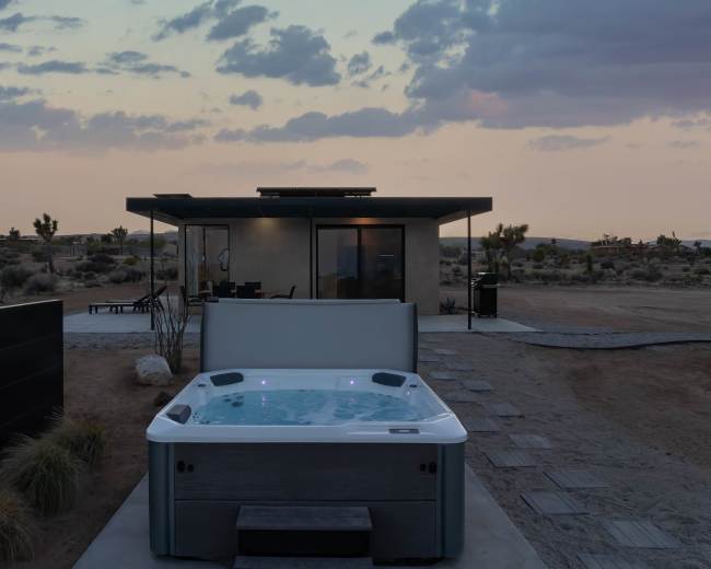A modern hot tub sits in the foreground with a desert landscape and a minimalist house in the background under a cloudy sky.