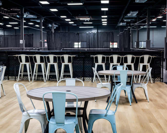 The image shows a spacious indoor venue with several round tables and metal chairs, with a backdrop of a netting area and ample overhead lighting.