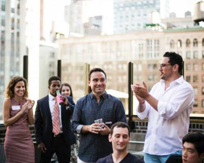 A group of people gathers on a rooftop, with some applauding and others smiling, in an urban setting.