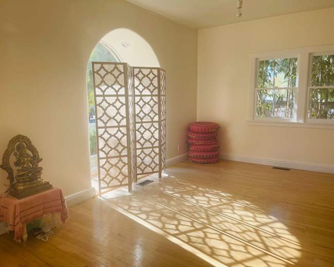 The image shows a bright, minimalist room with light-colored walls, a decorative screen casting patterns on the floor, and a small altar with a statue in the corner.