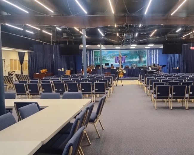 The image shows a spacious indoor setting with rows of blue chairs facing a stage, adorned with a large mural and equipped for a gathering or worship service.
