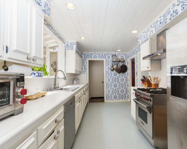 The image shows a narrow kitchen featuring white cabinetry, a blue and white tiled backsplash, and stainless steel appliances.