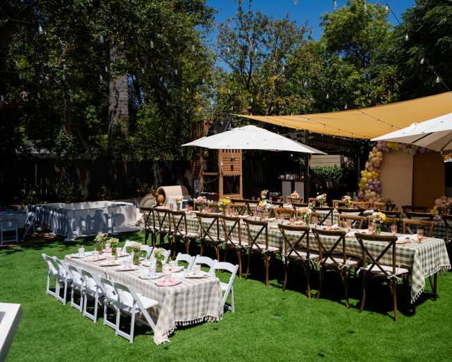 A backyard party setup features long dining tables covered with checkered tablecloths, surrounded by chairs, and shaded by large umbrellas under a sunny sky.