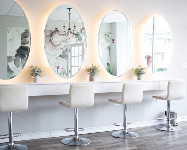 A modern vanity area with four white bar stools positioned in front of large oval mirrors illuminated by soft lights.