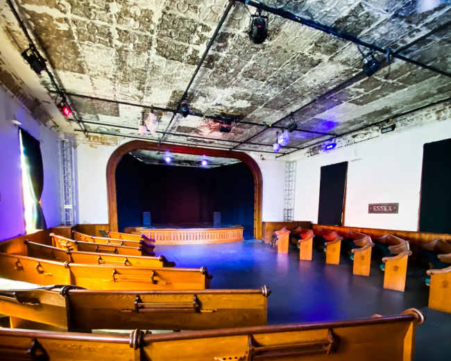 A theater interior with wooden seating arranged in rows facing a stage at the front, under a textured ceiling with colorful lighting.