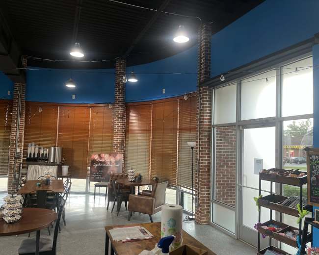 The image shows a modern café interior with brick walls, blue accents, wooden blinds, and several tables and chairs arranged in a well-lit space.