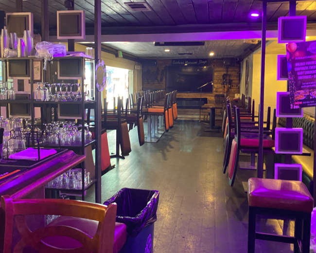 The image shows an empty bar area with tables and chairs stacked upside down, illuminated by purple lights.