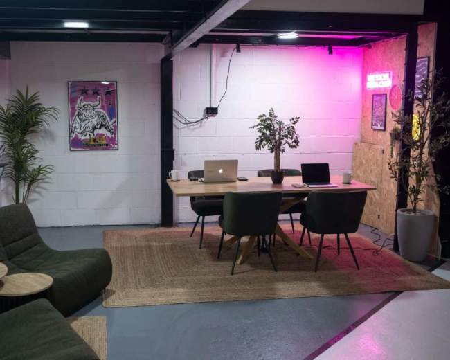 The image shows a modern office space featuring a table with laptops, chairs, and decor, surrounded by plants and artwork on the walls.