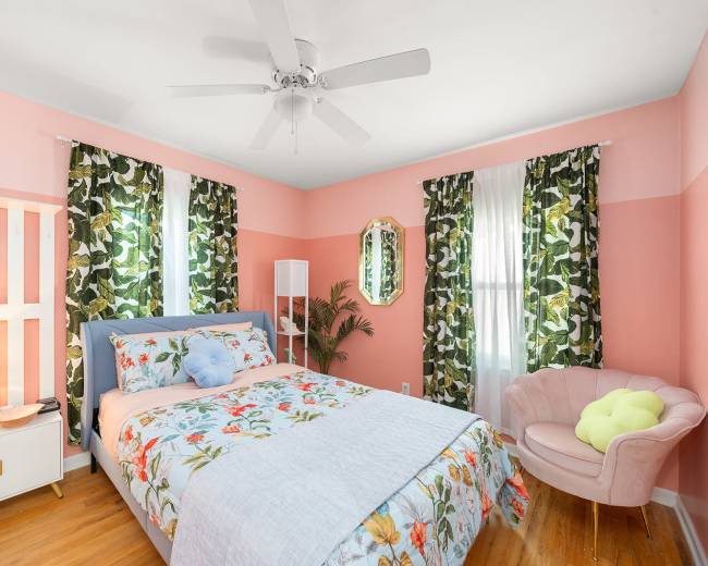 The bedroom features a bed with floral bedding, a pink accent wall, and large green plant-patterned curtains.