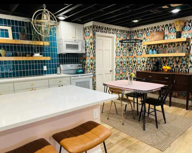 The image shows a modern kitchen with teal and patterned wallpaper, wooden shelves, and a dining table surrounded by black chairs.