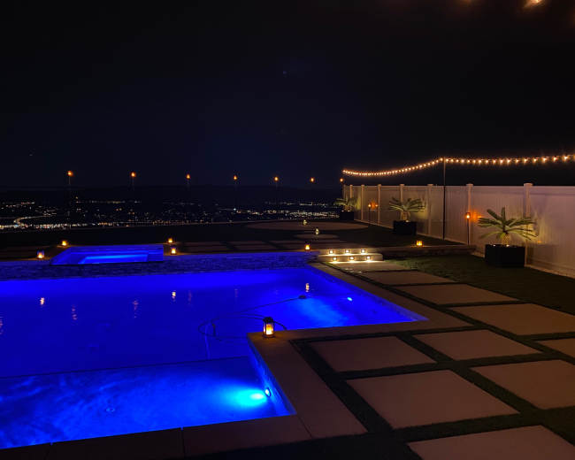 The image shows a nighttime view of a pool area with blue lighting and string lights illuminating the surrounding patio and landscape.