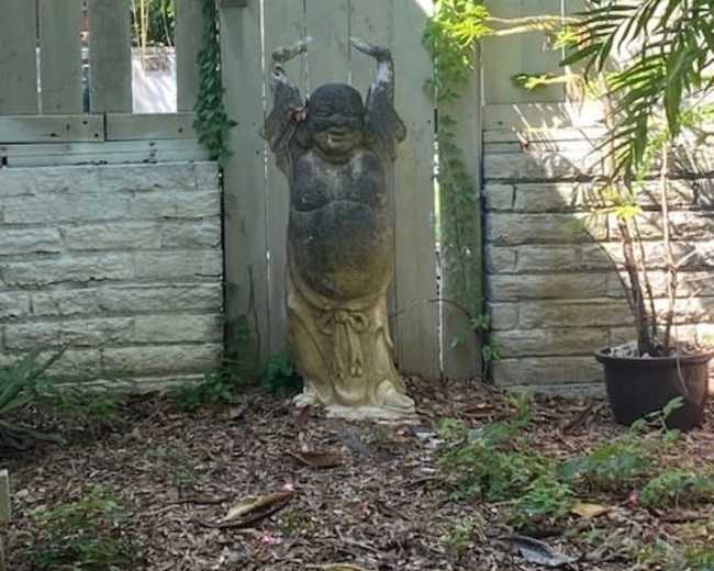 A stone statue of a smiling figure with raised arms stands beside a wooden fence in a garden with tropical plants.