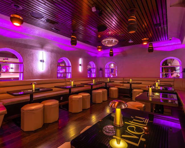 The image shows a dimly lit lounge with purple lighting, featuring plush seating areas, low tables, and decorative elements.