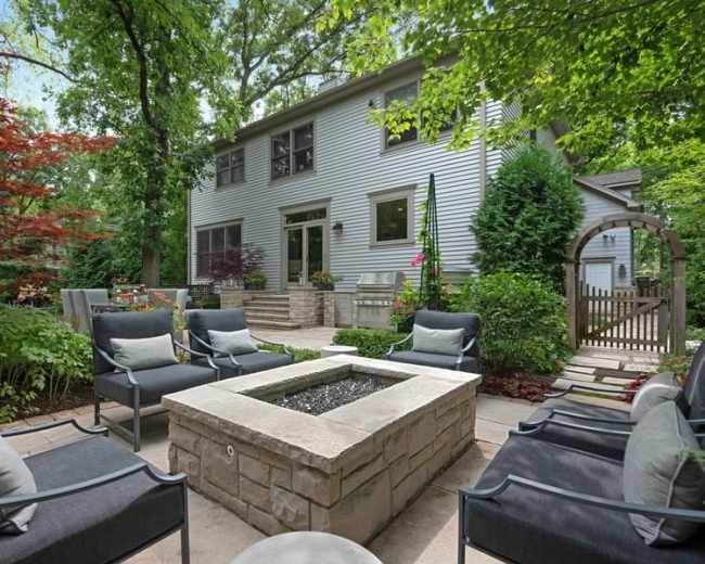 A stone fire pit is surrounded by gray chairs in a landscaped backyard with a two-story house and lush greenery.