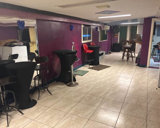 The image shows a narrow room with tiled flooring featuring bar-height tables and chairs, a red chair, and multiple windows providing natural light.