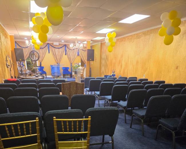 The image shows a room arranged with rows of chairs facing a stage, decorated with yellow balloons and drapery.