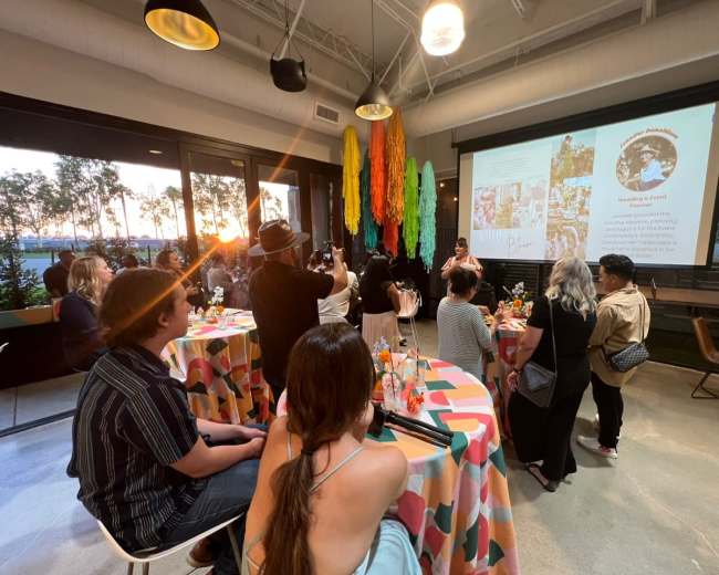 A group of people is attending an event in a brightly colored venue with decorative streamers and a presentation screen, while sunset light streams in through large windows.