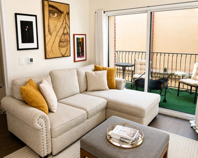 The image shows a cozy living room with a light-colored sectional sofa, a coffee table, and large sliding glass doors leading to a balcony.