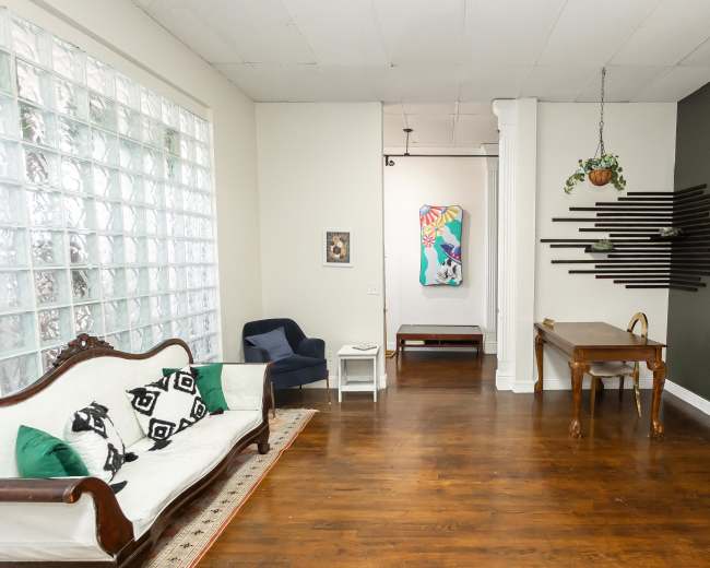 The scene shows a spacious room with a glass block wall on one side, a vintage sofa, a blue armchair, and a wooden dining table against a dark green wall, with artwork displayed in the background.