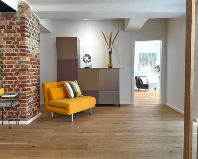 A modern living space features a yellow accent chair, a multicolored cabinet, and a brick wall, with natural light illuminating the hardwood floor.