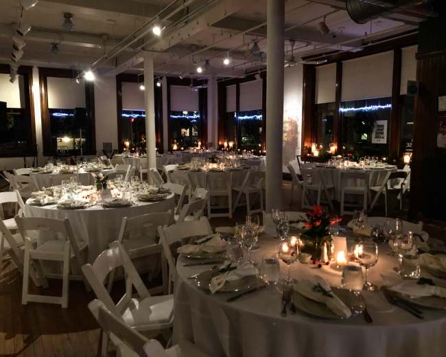 An elegantly arranged dining area with round tables set for a formal event, featuring white tablecloths, glassware, and centerpieces, illuminated by candlelight.