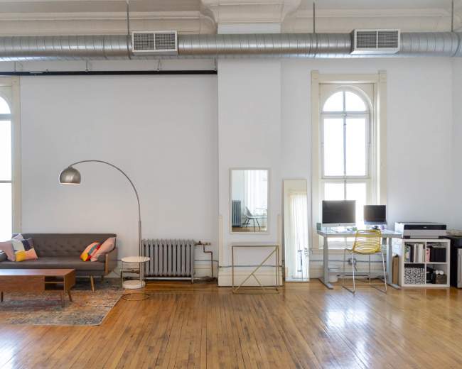 The image shows a spacious living area with a gray sofa, wooden coffee table, and desk setup, featuring large windows and wooden flooring.