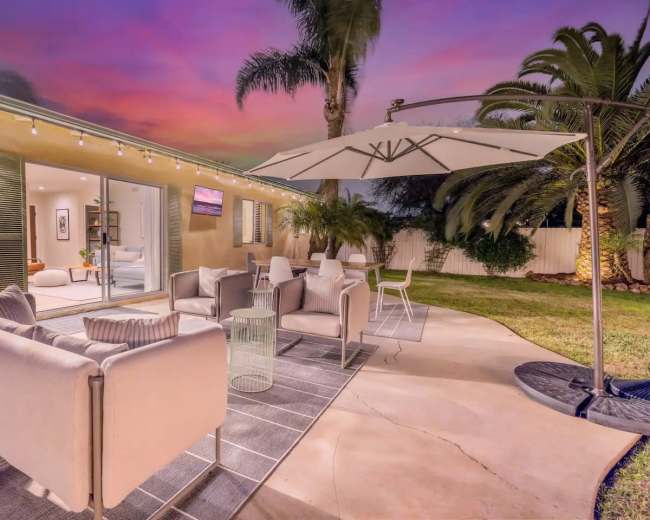A cozy patio features modern seating, an umbrella, and vibrant palm trees under a colorful sunset sky.