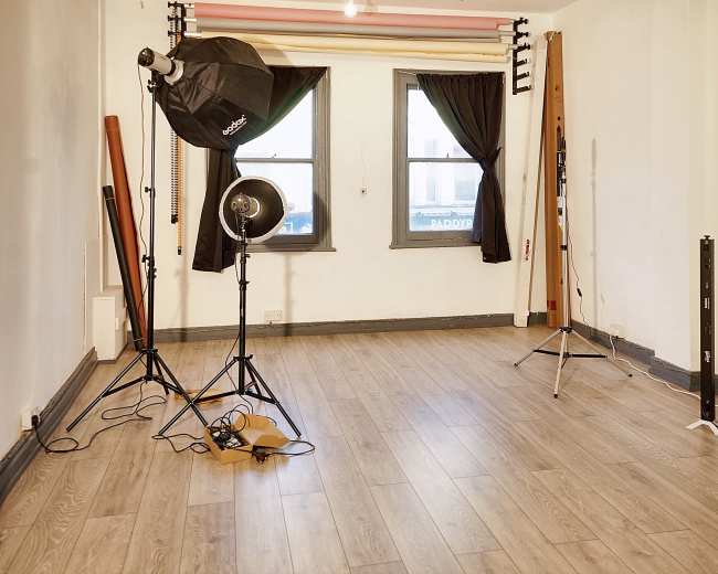 The image shows an empty room with wooden flooring, two windows covered with black curtains, and various photography equipment, including a light umbrella and stands.