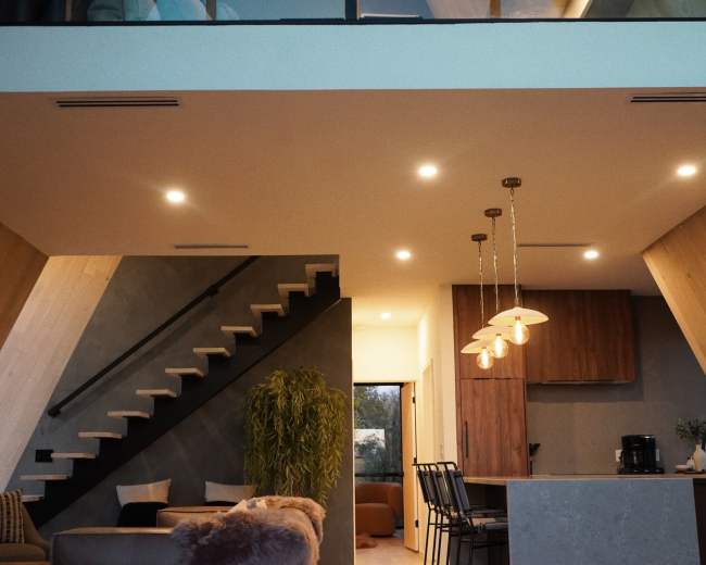 The image shows a modern interior of a two-story home featuring a staircase, a living area with a sofa, and a kitchen with pendant lighting.