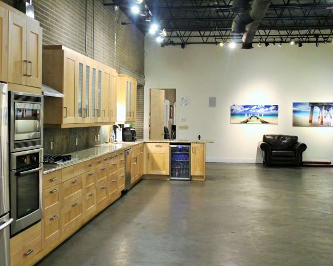 A modern kitchen with wooden cabinets and stainless steel appliances, set in an open space that includes a seating area and artwork on the wall.