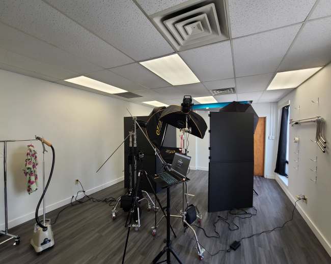 A photography studio setup with lighting equipment, a laptop, and dark backdrops in a spacious room.