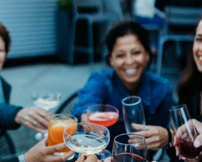 A group of people cheers with various drinks around a fire pit on an outdoor patio.
