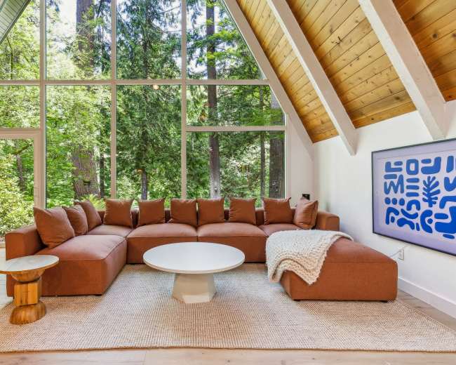 A spacious living room features a large sectional sofa, a circular coffee table, and a wall of windows overlooking a forested area.