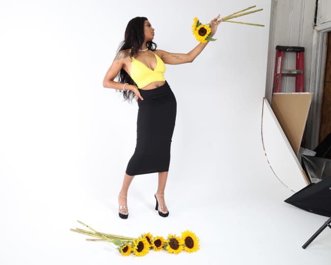 A woman in a yellow top and black skirt poses with sunflowers in a studio setting.