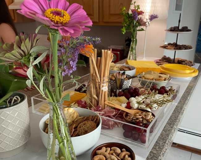 A variety of food items, including cheese, fruits, nuts, and snacks, is arranged on a kitchen counter alongside a decorative flower in a vase.