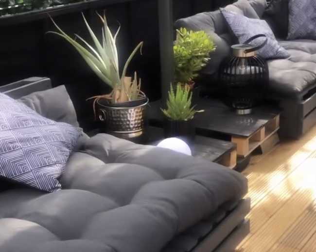 A cozy outdoor seating area featuring dark cushioned sofas, decorative plants, and accent pillows arranged on a wooden deck.