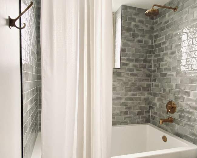 A bathroom featuring a bathtub with a white shower curtain, surrounded by gray tiled walls and brass fixtures.