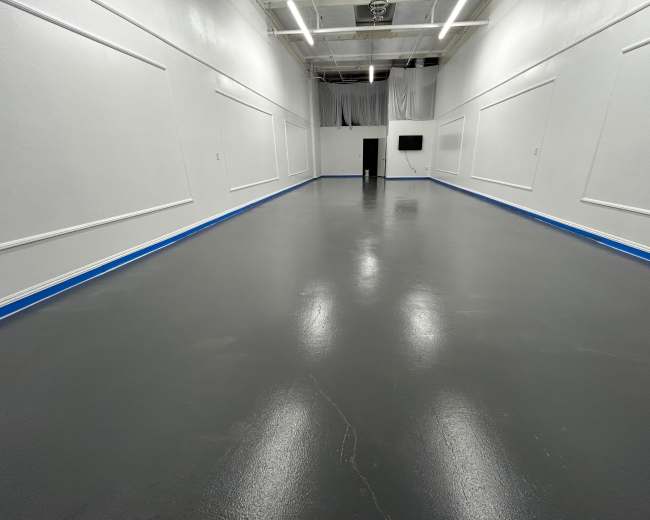 The image shows a spacious, empty room with a polished gray floor and white walls, featuring blue trim and a television mounted on one wall.