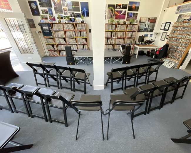 The image depicts a small performance space with rows of black folding chairs facing a stage, surrounded by walls lined with vinyl records and music memorabilia.
