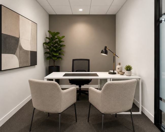 The image shows a modern office space with a white desk, two light-colored chairs, a small plant, and a framed abstract artwork on the wall.