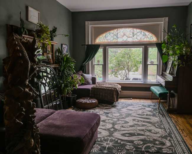 The room features a large window with stained glass, surrounded by plants, a purple couch, and a piano against the wall.