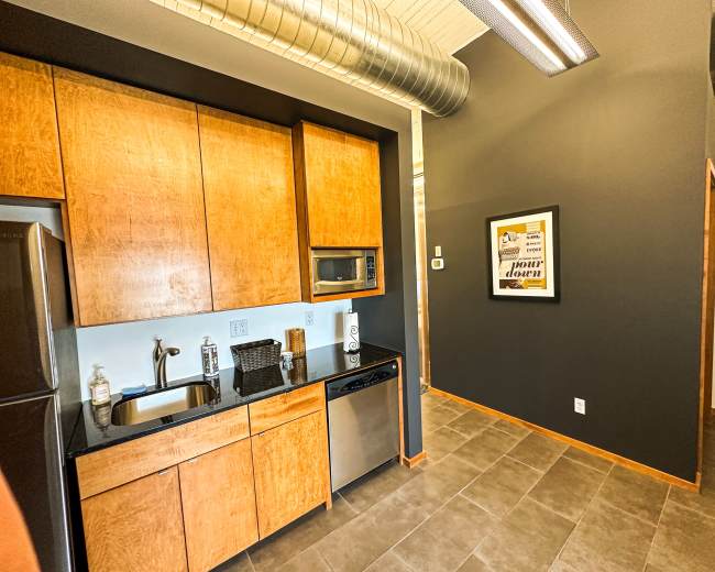 The image shows a modern kitchen area with wooden cabinetry, stainless steel appliances, and a dark accent wall.