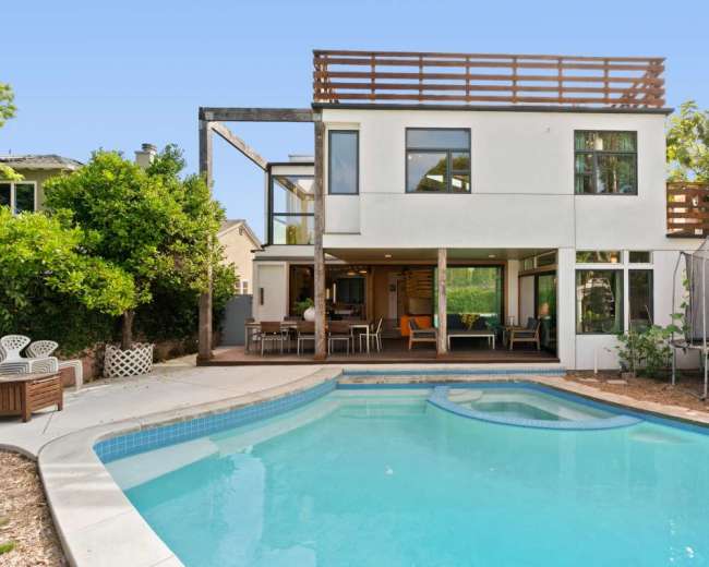 The image shows a modern two-story house with a pool in the backyard, featuring a spacious deck and a trampoline.