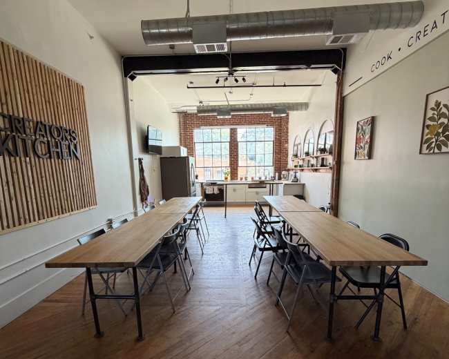 The image shows a spacious kitchen area with two long wooden tables set up for a gathering, illuminated by natural light from large windows.