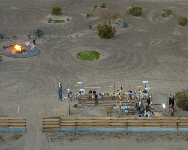 A gathering of people is set up around a long table under lights in a desert landscape, with a fire pit and various plants in the background.