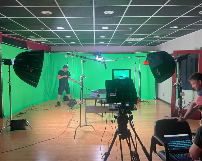 A video production studio is set up with green screens, lighting equipment, and a camera focused on a table.
