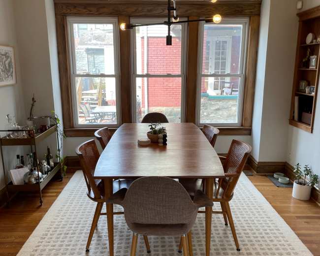 A dining room features a large wooden table surrounded by various chairs, illuminated by a modern light fixture, with windows showcasing an outdoor view.
