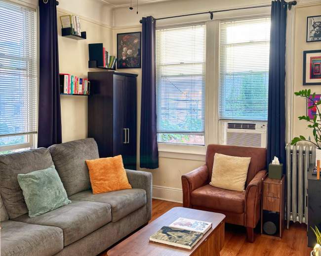 The image shows a cozy living room with a gray sofa, a brown armchair, a wooden coffee table, and large windows allowing natural light to enter the space.