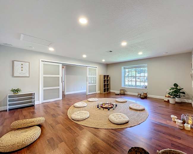The image shows a spacious, well-lit room with a circular arrangement of cushions around a decorative centerpiece on a large round rug, flanked by shelves and plants.