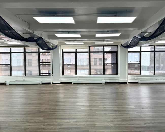 The image shows a spacious room with large windows, modern lighting, and a wooden floor, featuring sheer curtains draped from the ceiling.